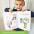 A customer photo of a smiling blonde boy holding up the I can Doodle Rhymes book with a green marker in his right hand. The left page of the open book shows a boy with ice cream dropped from his cone and the words "this lad is sad." Below is a girl with a 2 scoop ice cream cone with the text "she is glad." On the right is a boy with a cone and drawn in is a smile on his face and ice cream drawn in the cone with the text "can you make a sad lad glad?"
