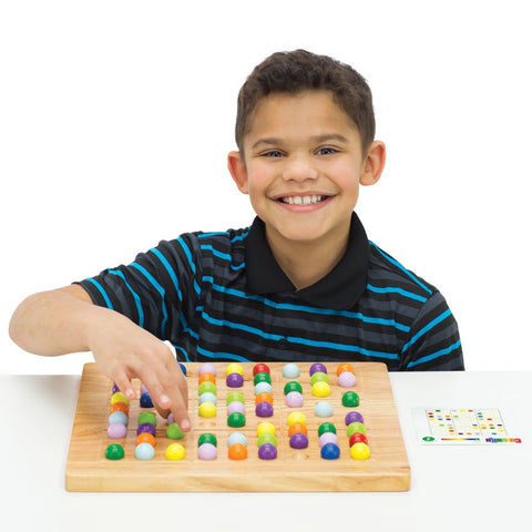 ColorKu is being played by a dark-haired boy in a striped shirt, smiling from ear to ear. He has his right hand over the game board pinching a lavender piece and putting it in place. The large wooden boar has spots cut out to put the wooden colored balls into place. There is a game card to the right of the board. The colored balls on the board are red, orange, yellow, bright green, dark green, light blue, dark blue, light purple, and dark purple.