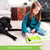 A customer photo of a young brunette girl playing with the Smart Farmer game next to her black dog on the floor. She is placing an animal piece on the board while looking at the instruction book off to her left. The green rectangle-shaped game board has white fence pieces all around the edge and a few through the middle. You can see a horse and 2 pigs inside the fence on the board. Off to the side are 2 cow pieces, and fence pieces.