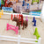 A close up of the Horse Academy game. The game board is a sand-colored rectangle with white fence pieces around the edges. There a several jumping fence pieces in many colors on the game board and a red girl character on top of a horse piece in between the fences. You can see the game box in the background, out of focus.