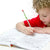 A blonde curly-haired boy in a red shirt is sitting at a white table and writing in the Daily 6 Trait Writing Grade 1 book. The book pages explain how to gather and plant seeds. The left page has 4 boxes to draw a sequence of gathering. The right page shows a tree with lines to answer questions below.