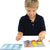 A young blonde boy is playing the Smart Cookies game. He is placing a blue square piece in the lower-left square of the game board with his left hand. The game board in front of him is filled with the red, yellow, and blue cookie pieces that are triangle, circle, and square shaped. To his upper-right is the spiral-bound game challenge book.