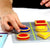 A close-up of a boys’ hands playing with the Smart Cookies game. He is placing a blue circle cookie piece with his right hand on the right-middle of the board. The game board in front of him is filled with the red, yellow, and blue cookie pieces that are triangle, circle, and square shaped. You can see the edge of the game challenge book to his upper-right.