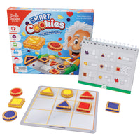 The Smart Cookies game box with the contents setup in front of it. The box shows an illustration of a man, looking like Albert Einstein, pulling a cookie sheet out of an oven with the cookie game pieces jumping off the board. To the right is the spiral-bound game book standing, showing a challenge page. In the front is the silver game board with a 3 by 3 grid printed on top. There are red, yellow, and, blueish-purple cookie playing pieces in triangles, circles, and squares on the board and off to the side.