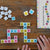 Candygrams game being played on a wood table. There are hands placing a letter in the crossword. The words spelled in the crossword are quest, at, hive, holiday, awaken, ham, and noun. There are tiles in the upper-left that are turned over, except 1 J tile. To the right are 2 large dice with colored dots on each side in pink, yellow, and blue. In the upper-right is the game box that shows the title in alternating colored tiles.