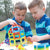 2 blonde boys in striped shirts are outside and are putting together a large shape, made up of the GeoSmart Educational Deluxe pieces on a picnic table. The pieces are geometrical shapes in squares and triangles and are a variety of different colors.  The object they are putting together is rounded and being held up by the boy on the left. The boy on the right is reaching out to grab a piece on the table. There are pieces in stacks and piles on the table.