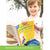 A customer photo of a young blonde boy holding the What Your Kindergartener Needs to Know book. He appears to have stopped reading to look and smile at the camera. The cover has a dark yellow background. The book title is written at the top in blues. Below and to the left is a child’s drawing page of 2 stick figures with a sun above them and a red K fridge magnet over the top of the drawing. Below is a photo of 4 children looking through a book in a classroom.