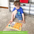A customer photo of a young blonde boy sitting on the carpet and placing his hand on the Bambino L U K controller that is on top of a game book. You can see the book through the controller and shapes on the book and tile, that you match up. The yellow tiles have a duck, apple, flower, heart, car, and house on them. The book under shows a watering can, clock, candy, teddy bear, teapot, and snowman.