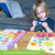A customer photo of a blonde girl sitting in front of a coffee table working on a Djeco So Pop Collages project. She is looking down at the dot she is holing in her hand and a glue stick in the other with her tongue out as she is concentrating. The project in front of her is a butterfly with flowers. There are many dots and shaped already glued onto the page. To the left is the project box with a giraffe on the cover.