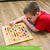 A customer photo of a brunette boy laying on the floor playing ColorKu. He is looking down at the board in front of him and holding a bright green piece in his right hand. The board is almost full with the colored ball pieces. The wooden ball pieces are red, orange, yellow, bright green, dark green, light blue, dark blue, light purple, and dark purple.