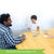 A customer photo of a father and son playing Tiny Polka dots at a round table. They are both smiling and reaching out to grab cards. There are many cards and card piles on top of the table, both face up and face down. The game box is standing up surrounded by the cards.