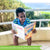 A customer photo of a, smiling, young black-haired boy sitting on a balcony near the ocean holding the Sharpeners Geography Grade 3 book. The book is open toward the boy and shows the cover to the camera. The book cover is mainly orange with a white top containing the title and 2 sample pages in the middle. To the right is a huge pencil illustration, standing from bottom to top. The back cover is white with images of several books from the publisher.