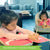 A customer photo of a young girl in a light pink shirt is sitting at a table and coloring in the Draw + Learn Animals + Places book, which is open to show a white cat shape on a pink page.  To the right is a toddler in a high chair, wearing a hot pink shirt and playing with wood blocks. The background is of a living room.